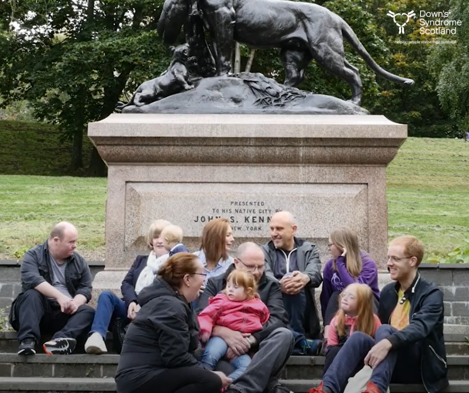 Families and carers sitting relaxing with their children at the based of a statue in a park. 